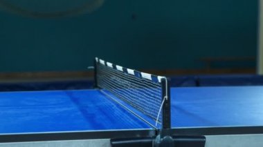 Çocuklar bir okul spor salonunda masa tenisi oynuyorlar. Masa tenisi maçını ve sarı topun hareketini masanın bir tarafından diğer tarafına hızlıca görebiliriz..