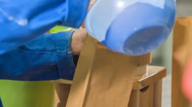 Genç bir çocuk yavaşça büyük mavi bir kaptan kahverengi bir kesekağıdına un döküyor..