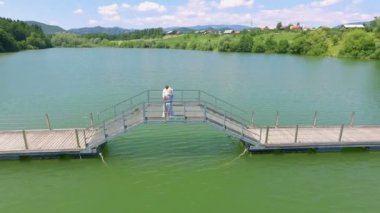 Bir aşk çifti gölün karşısına geçen bir köprünün tepesinde duruyor ve iyi vakit geçiriyorlar. Hava görüntüsü.