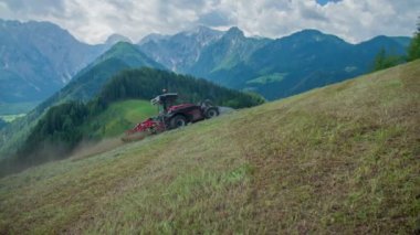 Bir traktör dik bir tepede saman hazırlarken saman etrafta uçuşuyor. Arkadaki dağlar güzel görünüyor..