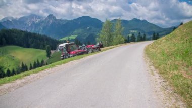 Küçük kırmızı bir traktör beton bir yolda geri gidiyor. Bir çiftçi dik bir tepede saman hazırlamayı bitirdi. Manzara çok güzel..