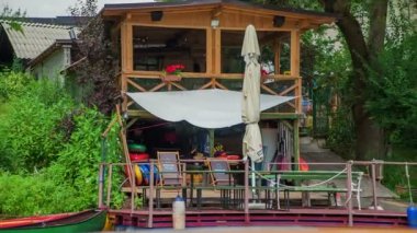 Bir tekne su üzerinde gidiyor ve nehir kıyısında küçük bir dükkanın önünden geçiyor. Yelken ve yüzme için her türlü ekipmanı satıyorlar..