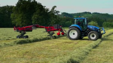 Mavi bir traktör birkaç farklı saman hattından geçiyor. Samanların çizgilere ayrıldığını görebiliyoruz..