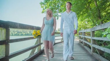 İki genç insan gölün etrafındaki ahşap bir patikada birlikte yürüyorlar. Bir kadın sarı bir buket tutuyor..