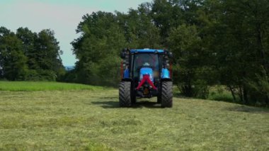 Büyük mavi bir traktör büyük bir çim tarlasına geliyor. Ona bağlı bir döner tırmık mekanizması var..