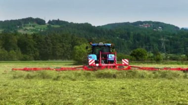 Mavi bir traktör tarladan geçiyor ve bir çiftçi saman hazırlıyor. Arka planda güzel tepeler var..