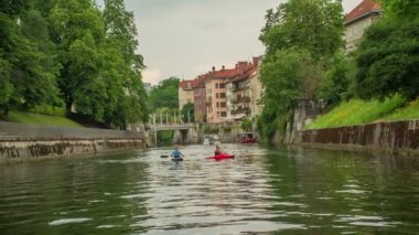 İki adam Ljubljanica nehrinde kano sürüyor. Nehrin her iki kıyısında da birçok ağaç var..