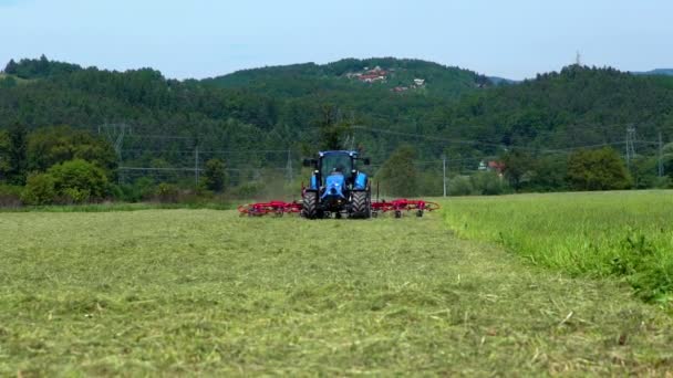 夏天的一天 一辆蓝色的大拖拉机正驶过田野 在农业机械的帮助下 农场主正在准备干草 — 图库视频影像