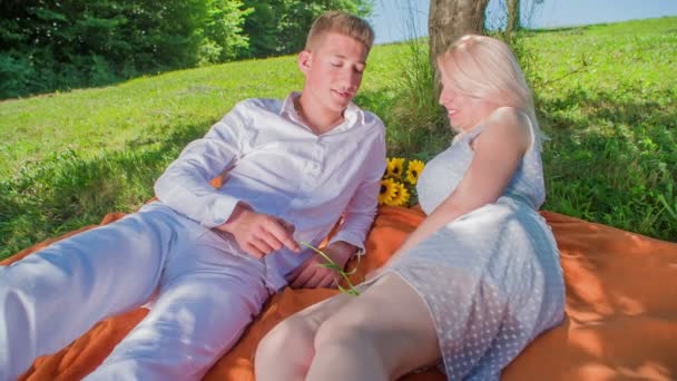 一对年轻夫妇躺在一棵树下的橙色毯子上 正在共度一段浪漫的时光 一个男人用草叶碰女人的腿 — 图库视频影像