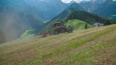 Traktörlü bir adam traktörünü yokuş yukarı sürüyor ve saman hazırlıyor. Tepe çok dik görünüyor..
