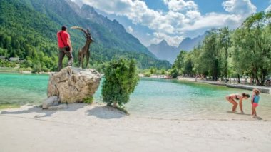 Bir adam büyük bir taşın üzerinde dağ keçisi heykelinin yanında duruyor ve gölde duran genç bir anne ve çocuğu var..