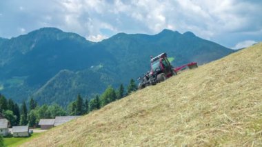 Genç bir çiftçi tepenin üzerinde araba sürerken ve saman hazırlarken arka planda dağlar muhteşemdir..
