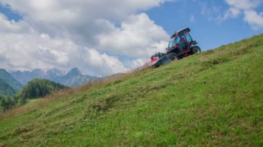 Bir traktör tepeden aşağı doğru gidiyor ve tarım makineleriyle çimleri kesiyor. Gün güzel ve hava güneşli..