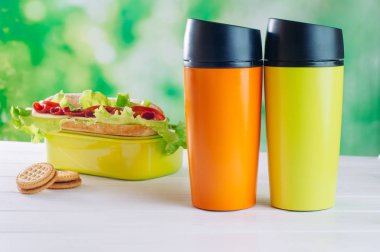 Beyaz ahşap arka planda, yemek kutusunun yanında renkli termos bardakları var.
