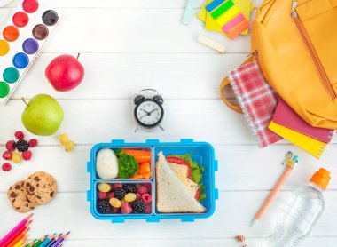 Sandviç, sebze, yumurta, taze böğürtlen ve saat ve okul aksesuarlarıyla birlikte beyaz ahşap arka planda açık büfe. Üst manzara, düz uzanma.