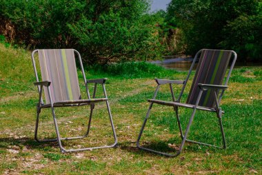 Nehir kıyısının yakınındaki çayırda yeşil ve gri çizgili bahçe sandalyeleri, seçici bir odak noktası.