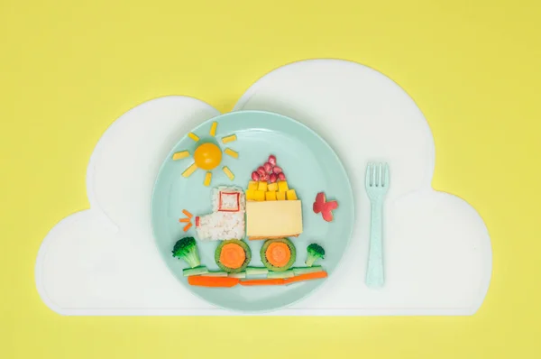 Kids food art concept: car from rice, sandwich and fresh fruits and vegetables on the plate with cloud napkin on the yellow background with blank space for text; top view, flat lay