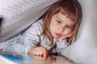 Yatak odasındaki battaniyenin altında kitap okuyan güzel küçük kız..