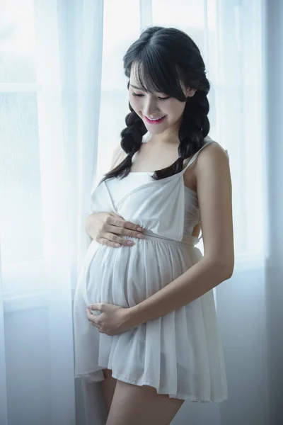 Young Pregnant Asian Woman Holds Her Hands on Her Swollen Belly, Love Concept. Pregnancy, Love, People and Expectation Concept - Happy Pregnant Woman Making Heart Gesture.