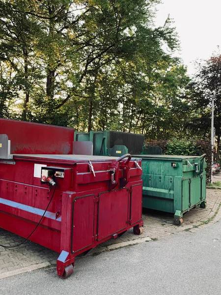 on an industrial site a green waste compactor and a red waste compactor stand side by side