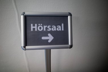 Beyaz harfli gri bir tabelada, konferans salonunda.