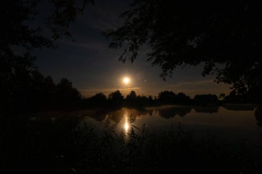 Bir gölün üzerinde yıldızlar ve ay gece gökyüzünde görülebilir