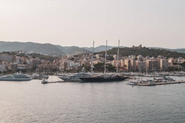 Palma de Mallorca limanının panoramik manzarası lüks yatlar, yelkenliler ve modern şehir binalarıyla dağlar ve Bellver Kalesi 'ne karşı. İspanya, Mallorca 'nın sahili, gün batımında sakin Akdeniz sularıyla