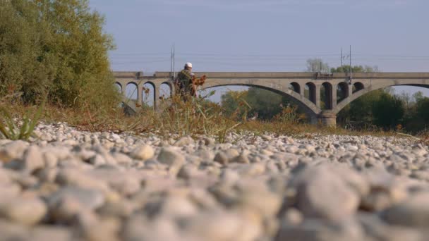 Рыбак идет к реке, а поезд пересекает мост. — стоковое видео