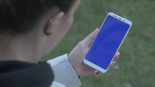 Девушка постукивает подержанными руками по синему экрану телефона, через плечо выстрел — стоковое видео