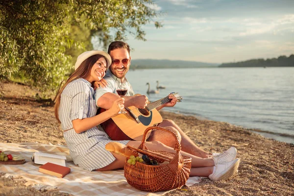 Pareja joven disfrutando de su tiempo, haciendo un romántico picnic en la playa. Tocar la guitarra y cantar — Foto de Stock