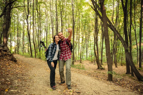 Amar a la joven pareja que usa camisas a cuadros tomando una selfie mientras camina por el bosque en un hermoso día de otoño. — Foto de Stock