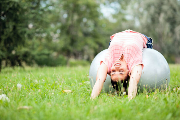 Beautiful young woman lying on her back on a fitness ball and stretching. Practicing pilates in city park