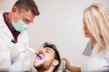 Olgun erkek diş hekimi ve diş Kliniği genç bir erkek hastada diş beyazlatma, Uv ışığı kullanarak güzel genç kadın asistanı