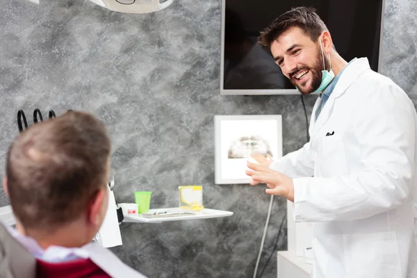 Odontólogo joven sonriente mostrando imágenes de rayos X a un paciente, consultando durante el examen en la clínica dental —  Fotos de Stock