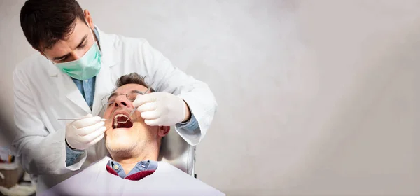 Jovem dentista do sexo masculino segurando um espelho dental e palheta dentária, realizando exame dentário em um paciente maduro do sexo masculino na clínica odontológica. Imagem de grande proporção com espaço de cópia no lado direito — Fotografia de Stock