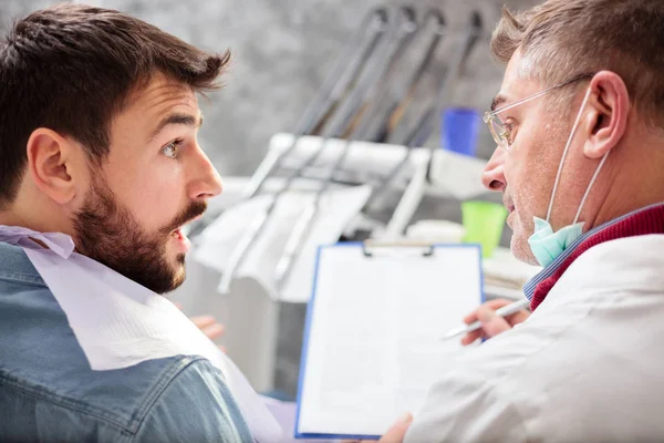 Dentista masculino maduro escribiendo los detalles del paciente en un portapapeles, consultando durante el examen en la clínica dental —  Fotos de Stock