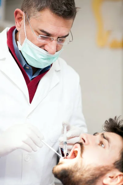 Dentista masculino maduro sosteniendo una jeringa y dando anestésicos al paciente masculino —  Fotos de Stock