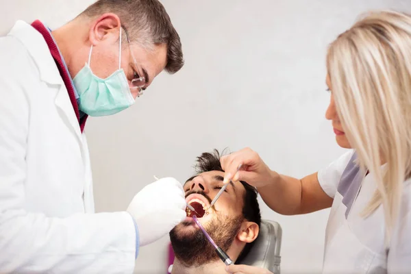 Dentista masculino maduro y hermosa asistente joven que realiza un examen dental en un paciente masculino joven en la clínica dental —  Fotos de Stock