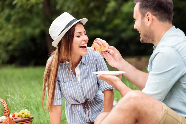 Feliz pareja joven tomando un desayuno relajante en el parque, comiendo croissants y tomando café — Foto de Stock