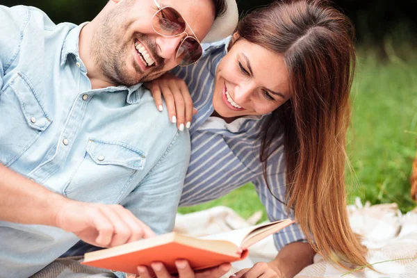 Pareja joven y feliz disfrutando del día en la naturaleza, leyendo un libro y tumbada en una manta de picnic — Foto de Stock