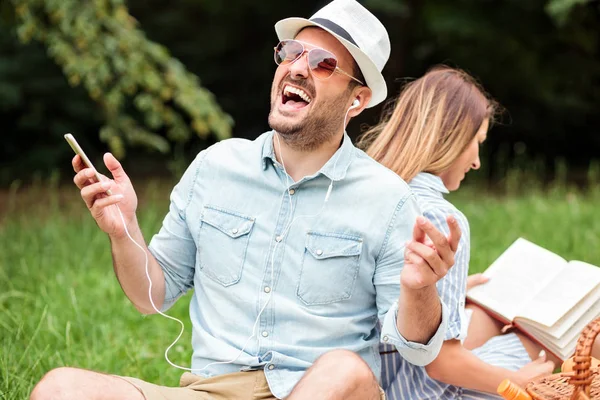 Joven disfrutando escuchando su canción favorita. Sentado espalda con espalda con su novia que está leyendo un libro — Foto de Stock