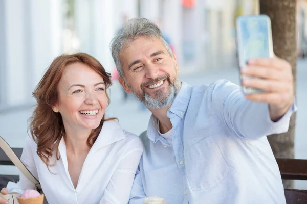 Mutlu olgun turistler çift bir bankta oturan ve selfie alarak. Karı koca selfie için poz