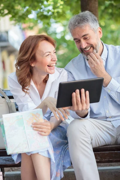 Mutlu gülümseyen olgun turistler bir tablet bakarak bir bankta oturan ve gülüyor. Kocası bir tablet tutuyor ve karısı bir şehir haritası tutuyor
