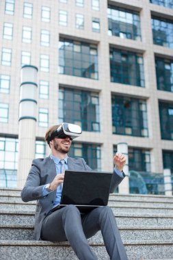 Genç işadamı VR gözlük kullanarak ve el jestleri yapma, bir ofis binasının önünde bir dizüstü bilgisayar üzerinde çalışıyor