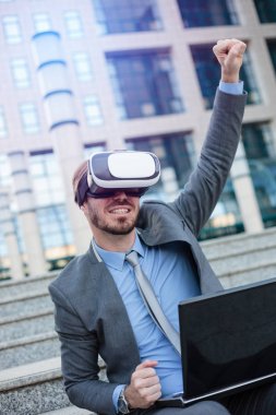 Bir ofis binasının önünde oturan VR gözlük kullanarak mutlu genç işadamı. Havaya yumruk yüksek başarı kutluyor