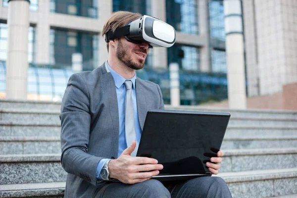 Successful young businessman using virtual reality simulator goggles and working on a laptop in front of an office building