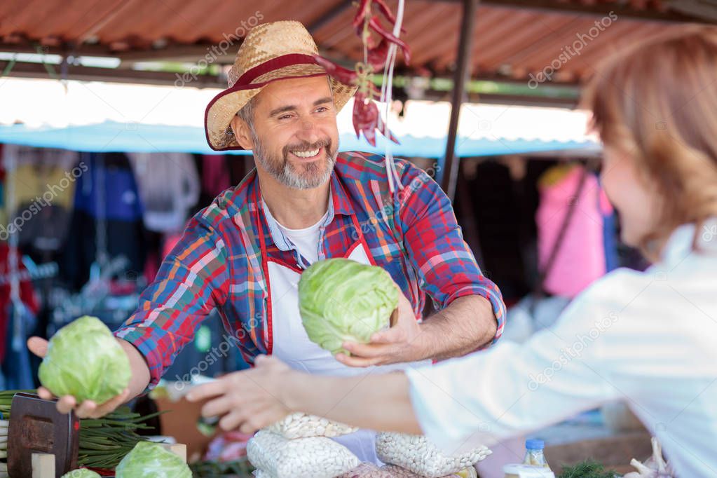 Happy smiling senior farmer standing behind the stall, selling organic vegetables in a marketplace