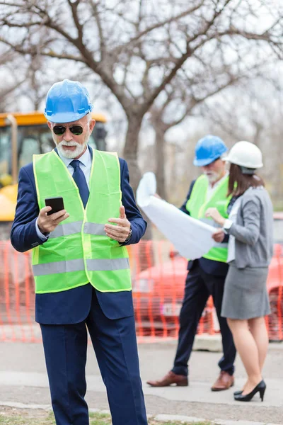 Ingeniero senior serio o hombre de negocios usando su teléfono inteligente mientras inspecciona un sitio de construcción — Foto de Stock