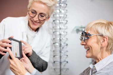 Smiling mature female ophthalmologist examining senior woman, holding an eye chart clipart