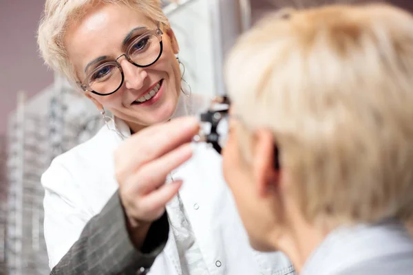 Mujer sonriente optometrista examinando a la mujer madura, determinando dioptría en clínica oftalmológica —  Fotos de Stock